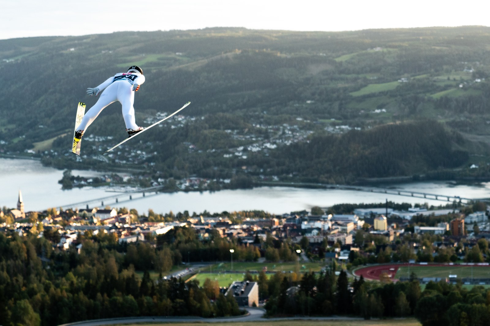 En skihopper i svevet med byen, vannet og skogkledde åser i bakgrunnen.