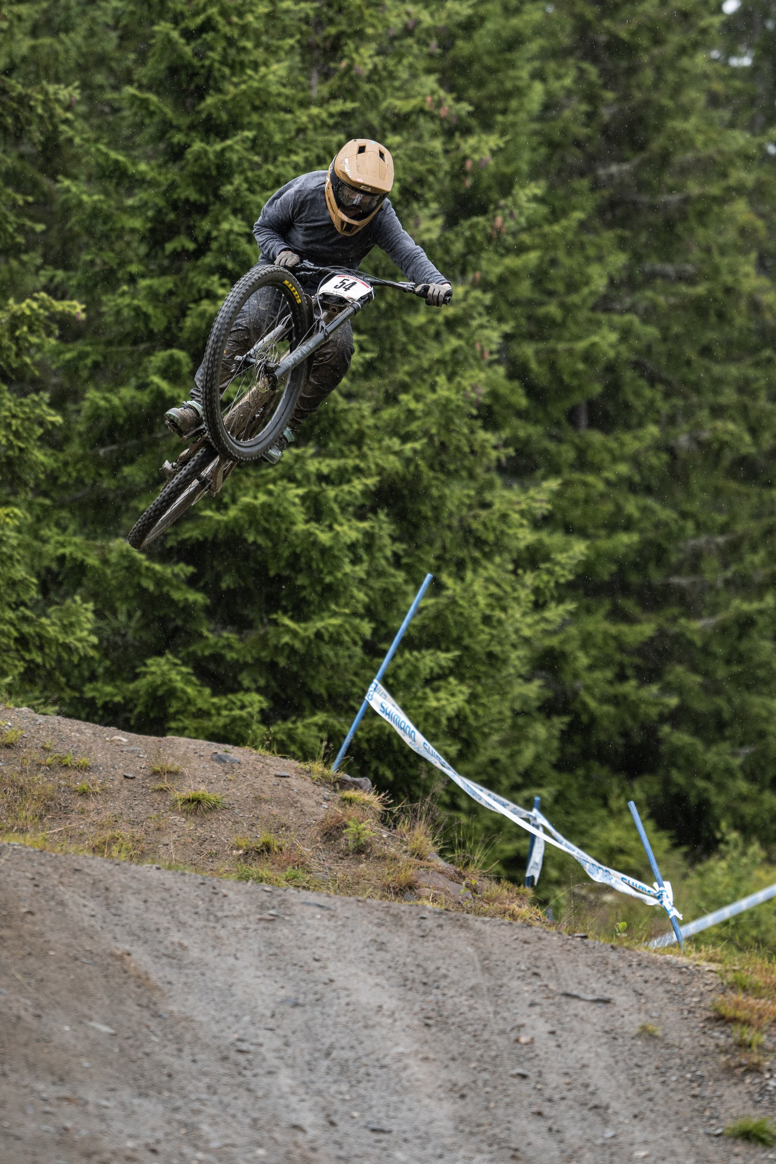 Terrengsyklist med lys hjelm legger sykkelen sidelengs i et hopp foran granskogen.